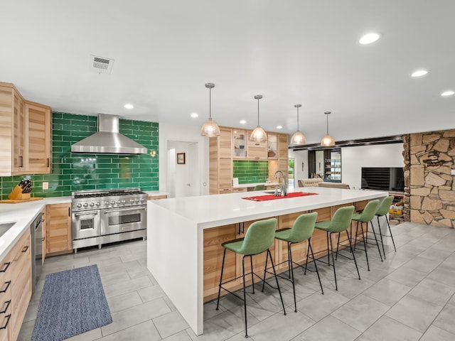 kitchen with a large island with sink, wall chimney range hood, light brown cabinetry, appliances with stainless steel finishes, and decorative light fixtures