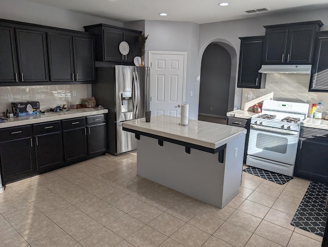 kitchen with visible vents, white gas range, dark cabinetry, under cabinet range hood, and stainless steel refrigerator with ice dispenser