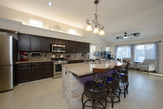 kitchen featuring pendant lighting, stainless steel appliances, tasteful backsplash, an island with sink, and ceiling fan with notable chandelier