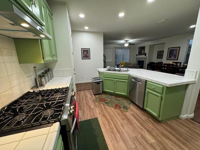 kitchen with exhaust hood, green cabinetry, light wood-type flooring, tile counters, and stainless steel appliances