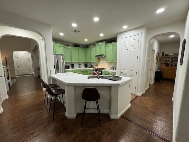 kitchen featuring tile countertops, a center island with sink, green cabinetry, stainless steel fridge, and dark hardwood / wood-style flooring