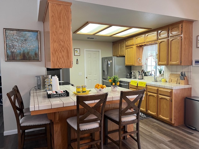 kitchen with tile countertops, a breakfast bar area, a peninsula, appliances with stainless steel finishes, and decorative backsplash