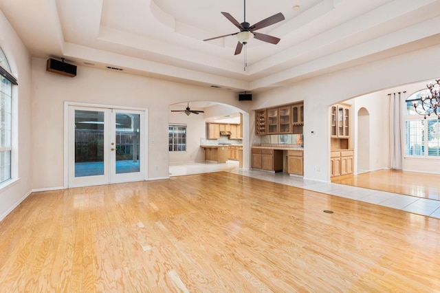 unfurnished living room with french doors, ceiling fan with notable chandelier, light hardwood / wood-style flooring, and a raised ceiling