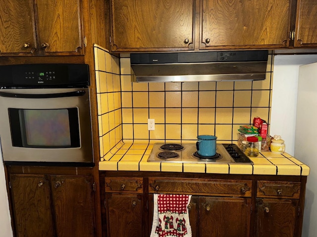 kitchen with tile countertops, oven, under cabinet range hood, stovetop, and tasteful backsplash