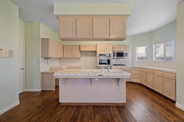 kitchen featuring appliances with stainless steel finishes, light brown cabinets, tile counters, and a kitchen island with sink