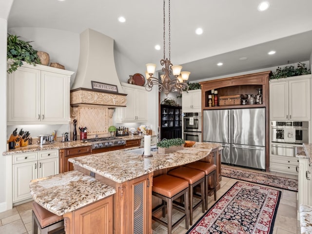 kitchen with custom exhaust hood, a kitchen island with sink, white cabinets, appliances with stainless steel finishes, and light stone counters