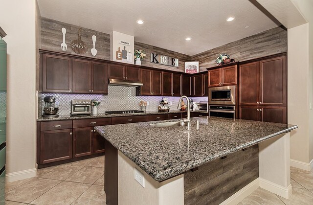 kitchen with dark stone counters, a center island with sink, sink, light tile patterned floors, and appliances with stainless steel finishes