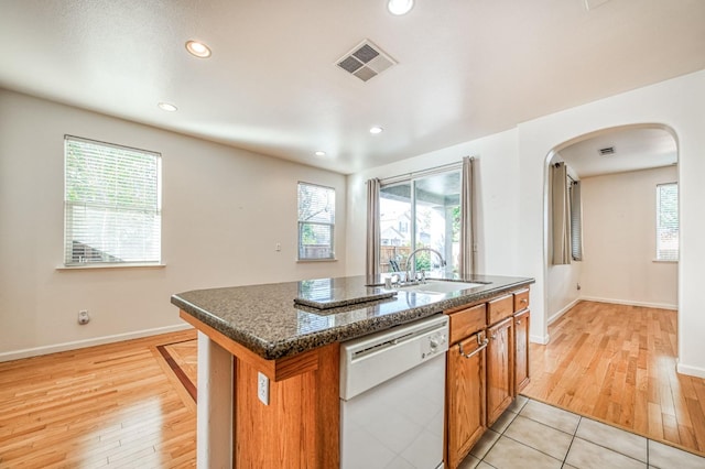 kitchen with sink, dark stone countertops, light hardwood / wood-style flooring, an island with sink, and white dishwasher