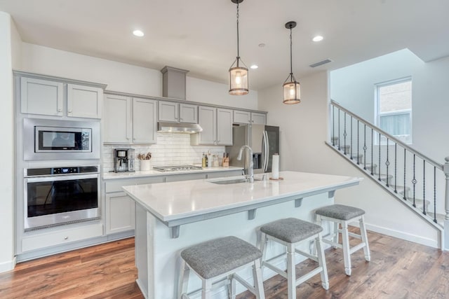 kitchen featuring decorative backsplash, decorative light fixtures, gray cabinets, a kitchen island with sink, and appliances with stainless steel finishes