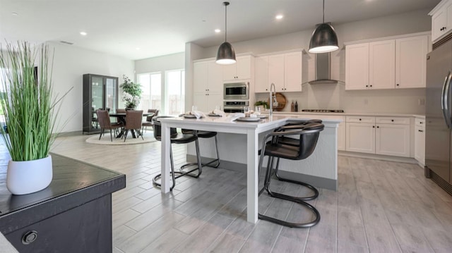 kitchen with wall chimney exhaust hood, stainless steel appliances, a kitchen island with sink, decorative light fixtures, and white cabinetry