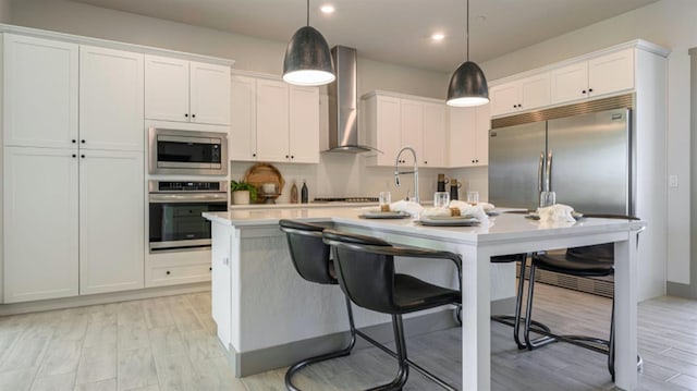 kitchen with wall chimney exhaust hood, built in appliances, decorative light fixtures, a center island with sink, and white cabinets