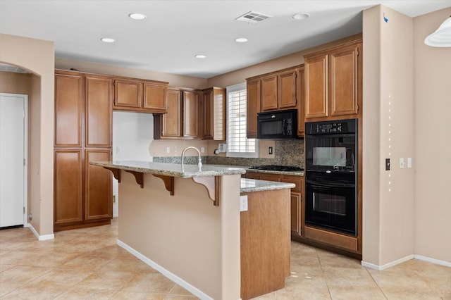 kitchen featuring light stone countertops, a kitchen breakfast bar, backsplash, a center island with sink, and black appliances