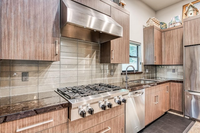 kitchen featuring appliances with stainless steel finishes, dark stone countertops, exhaust hood, decorative backsplash, and a sink