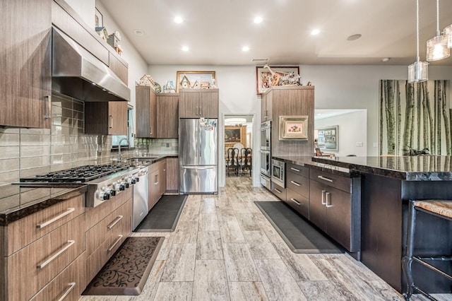 kitchen with pendant lighting, sink, range hood, appliances with stainless steel finishes, and a kitchen bar