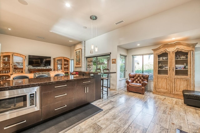 kitchen featuring a kitchen breakfast bar, dark brown cabinetry, pendant lighting, dark stone countertops, and stainless steel microwave