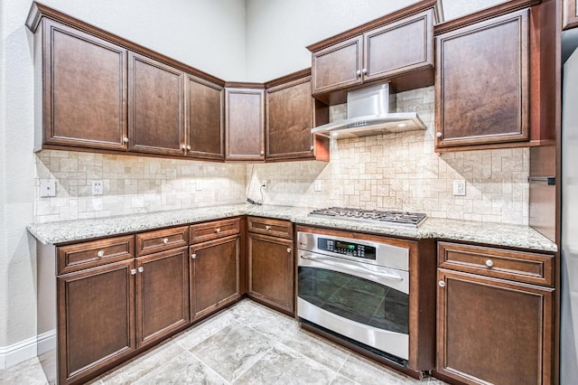 kitchen featuring backsplash, stainless steel appliances, light stone countertops, dark brown cabinets, and wall chimney exhaust hood