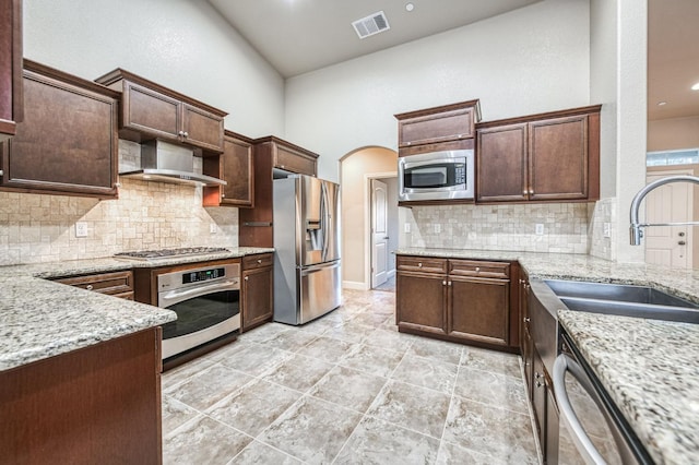 kitchen with wall chimney exhaust hood, sink, stainless steel appliances, light stone countertops, and a high ceiling