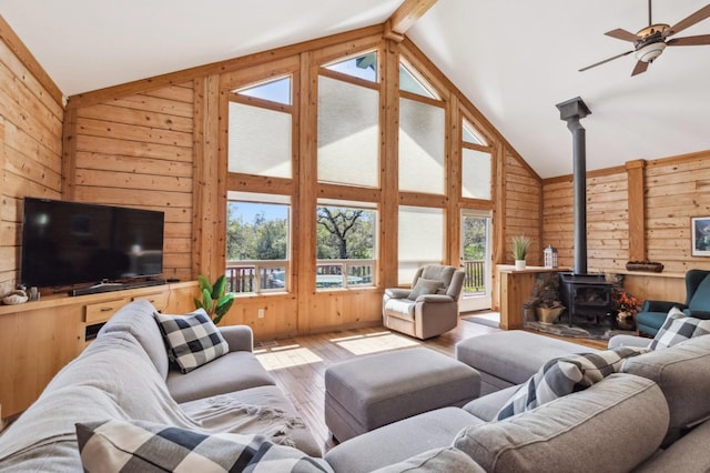 living room featuring wood walls, a wood stove, light hardwood / wood-style flooring, ceiling fan, and beam ceiling