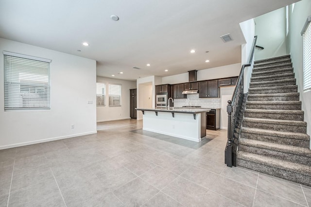 kitchen with stainless steel microwave, tasteful backsplash, a kitchen island with sink, a breakfast bar, and dark brown cabinets