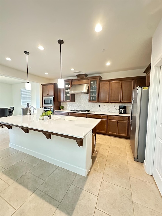 kitchen with a large island with sink, wall chimney range hood, hanging light fixtures, appliances with stainless steel finishes, and a kitchen bar