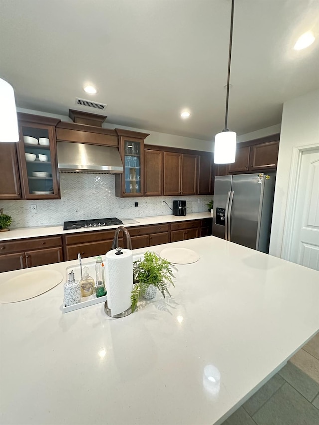 kitchen with stainless steel refrigerator with ice dispenser, dark brown cabinets, hanging light fixtures, and wall chimney range hood