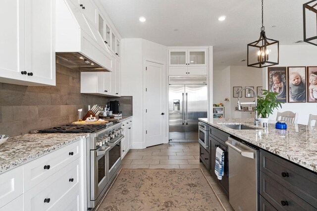 kitchen with custom exhaust hood, hanging light fixtures, built in appliances, sink, and white cabinetry