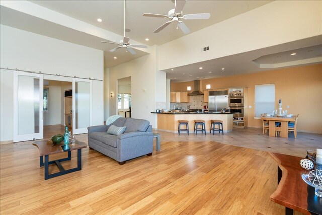 living room with a high ceiling, ceiling fan, a barn door, and light hardwood / wood-style flooring