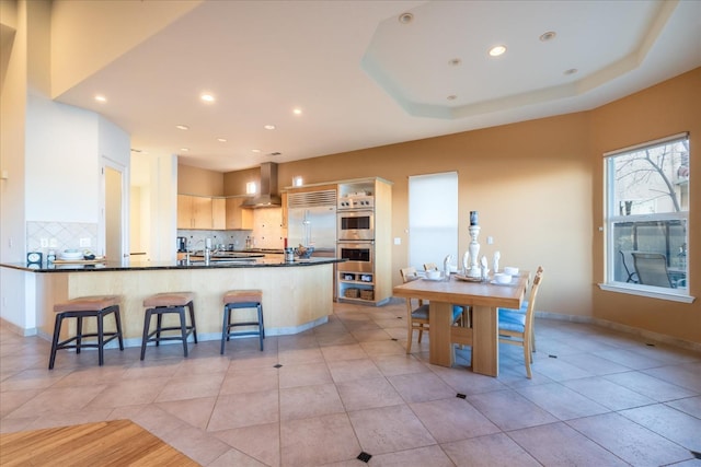 kitchen featuring stainless steel appliances, a raised ceiling, a breakfast bar area, backsplash, and wall chimney range hood