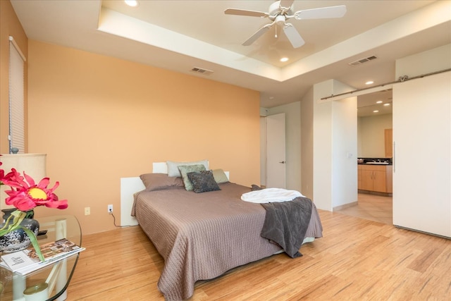 bedroom with a tray ceiling, a barn door, ensuite bathroom, and light hardwood / wood-style flooring