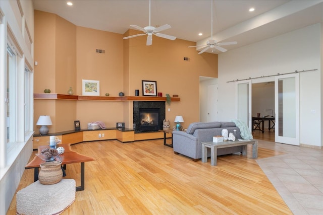 living room featuring a tile fireplace, a high ceiling, ceiling fan, a barn door, and light hardwood / wood-style flooring