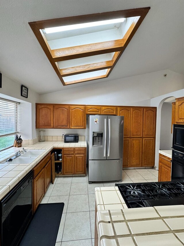 kitchen featuring tile counters, brown cabinetry, vaulted ceiling, black appliances, and light tile patterned flooring