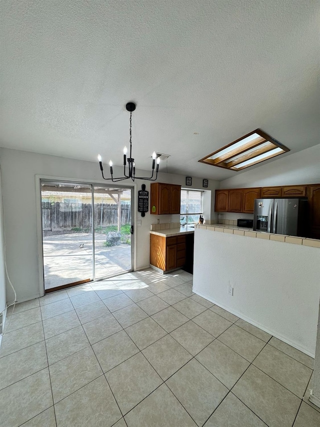 kitchen featuring stainless steel refrigerator with ice dispenser, tile counters, brown cabinetry, light tile patterned flooring, and a chandelier