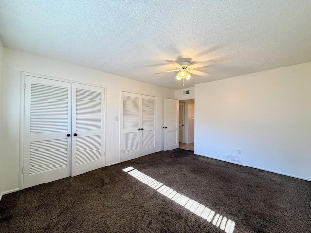 unfurnished bedroom featuring two closets, a textured ceiling, dark colored carpet, and ceiling fan