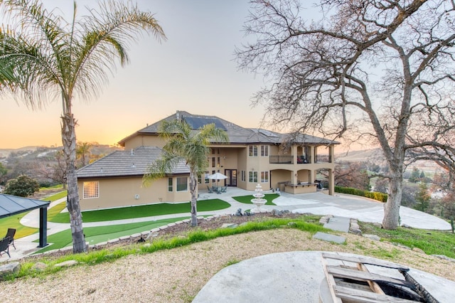 back house at dusk featuring a patio area, a lawn, and a balcony