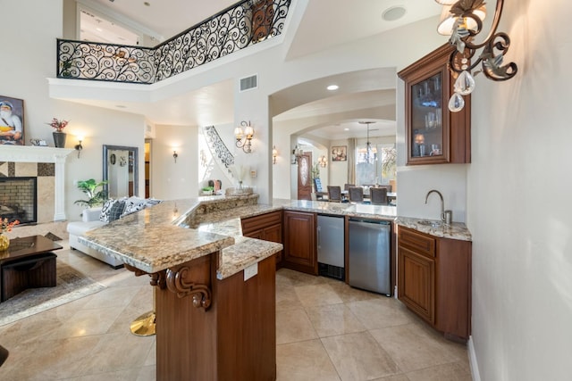 kitchen with dishwasher, kitchen peninsula, a towering ceiling, a tiled fireplace, and a breakfast bar area