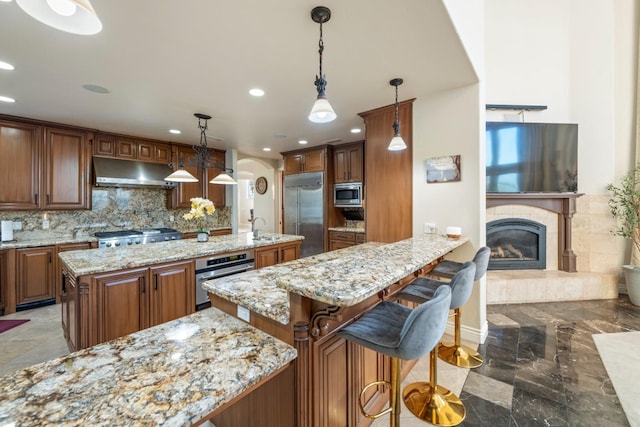 kitchen with built in appliances, decorative backsplash, hanging light fixtures, a kitchen island with sink, and a tile fireplace