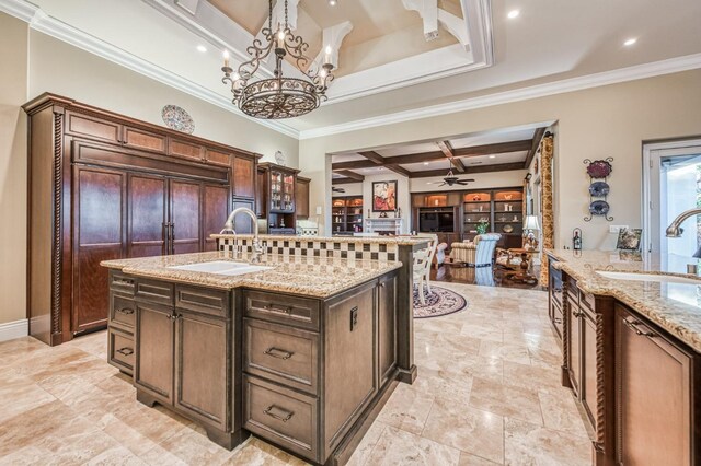 kitchen featuring coffered ceiling, an island with sink, ornamental molding, ceiling fan with notable chandelier, and beamed ceiling