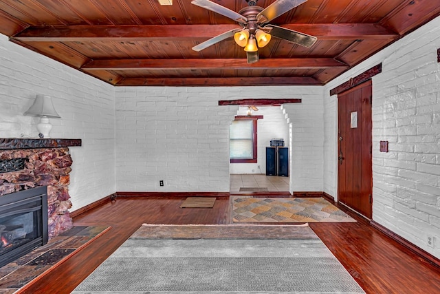 unfurnished living room featuring brick wall, dark wood-type flooring, a stone fireplace, ceiling fan, and wooden ceiling