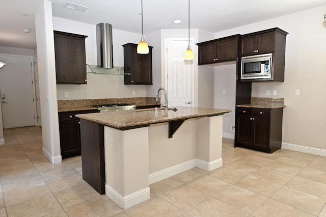 kitchen with a center island with sink, stainless steel microwave, decorative light fixtures, wall chimney range hood, and sink