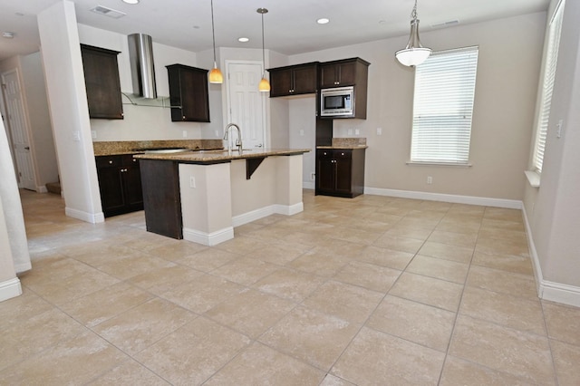 kitchen with a center island with sink, stainless steel microwave, decorative light fixtures, light stone countertops, and wall chimney exhaust hood