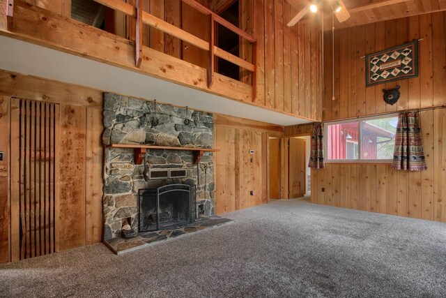 unfurnished living room featuring carpet, a towering ceiling, a stone fireplace, wood walls, and ceiling fan