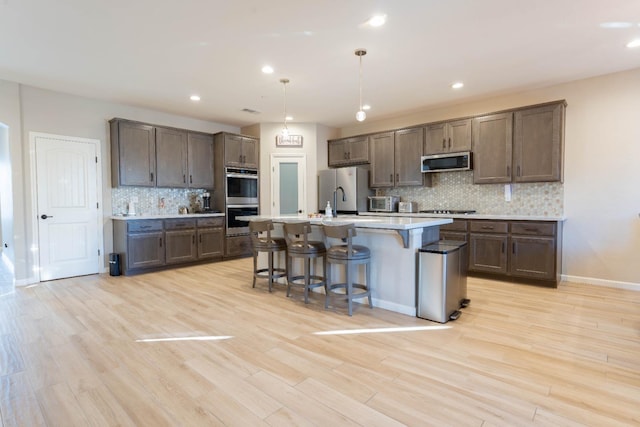 kitchen featuring stainless steel appliances, tasteful backsplash, an island with sink, hanging light fixtures, and light wood-type flooring