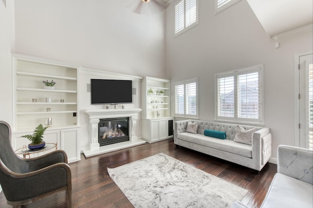 living room with ceiling fan, dark wood-type flooring, a towering ceiling, built in features, and a glass covered fireplace