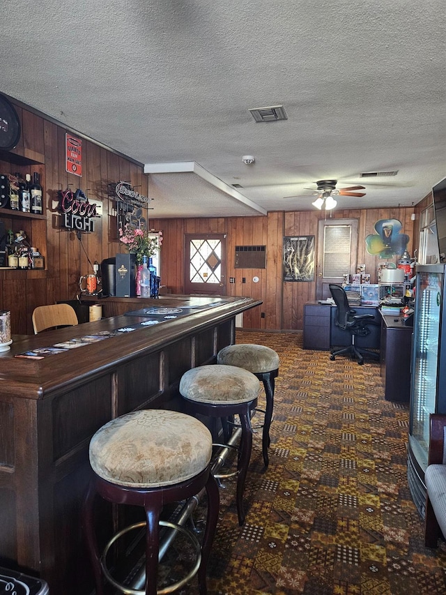 bar with dark colored carpet, ceiling fan, a textured ceiling, and wooden walls