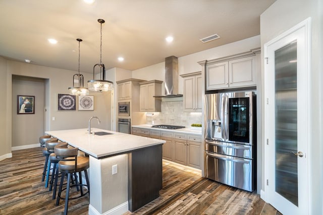 kitchen with appliances with stainless steel finishes, wall chimney range hood, sink, hanging light fixtures, and a center island with sink