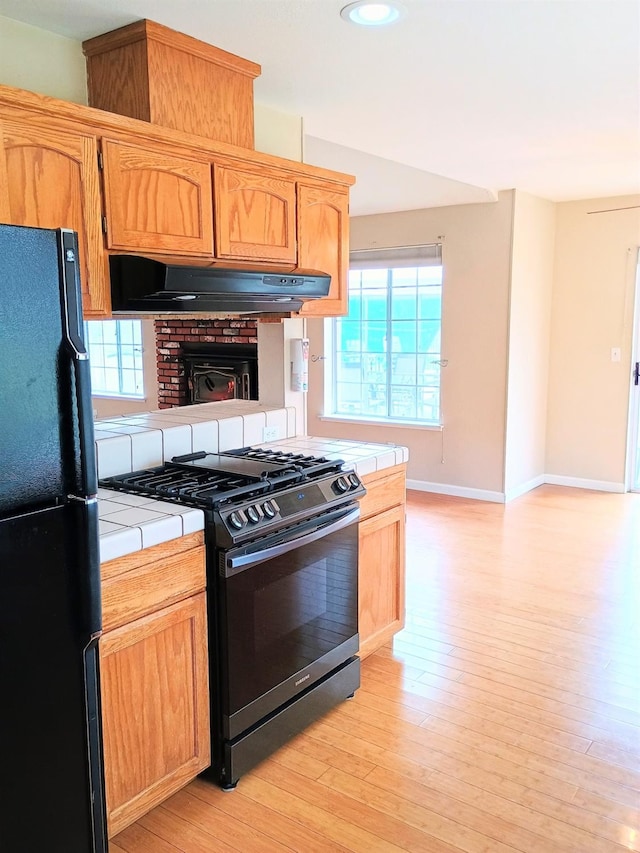 kitchen with stainless steel gas stove, ventilation hood, light hardwood / wood-style flooring, black refrigerator, and tile counters