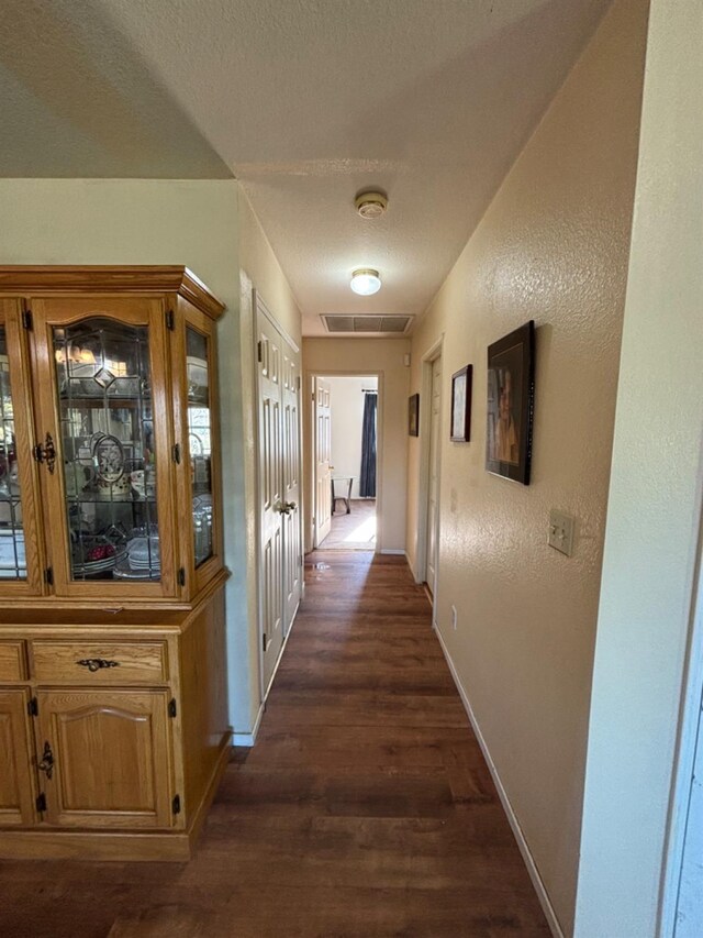 hall with a textured wall, dark wood-type flooring, visible vents, and baseboards