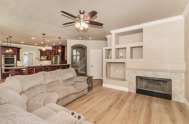 living room featuring crown molding, light wood-type flooring, a tile fireplace, built in features, and ceiling fan