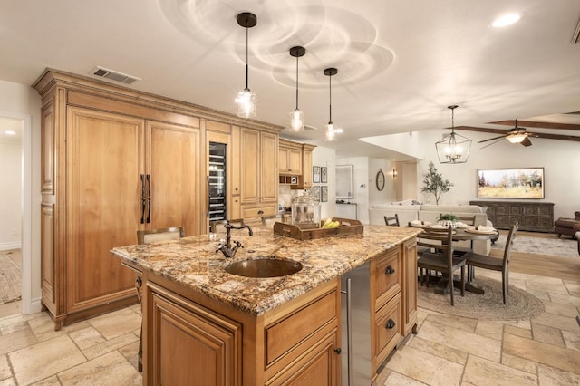 kitchen with sink, ceiling fan, light stone counters, an island with sink, and decorative light fixtures