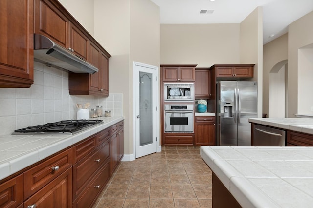 kitchen featuring light tile patterned floors, appliances with stainless steel finishes, backsplash, a high ceiling, and tile counters